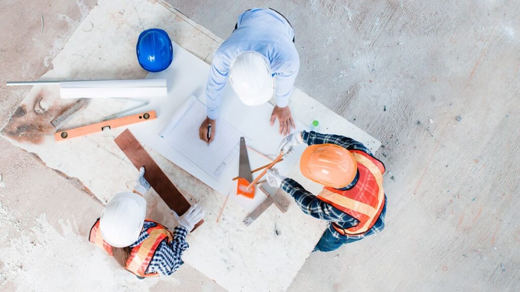 Three men standing looking at table with plans. Picture taken from above.